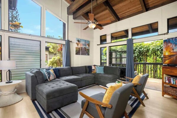 A spacious living room with a sectional sofa, a coffee table, and a fireplace. There are large windows and a balcony with a view of greenery.