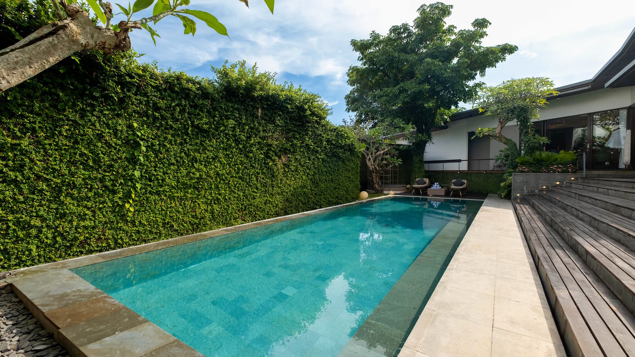 A rectangular swimming pool with clear blue water, surrounded by a stone border and a wooden deck.