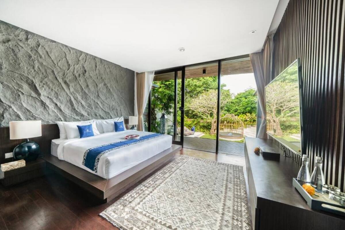 A modern bedroom with a large bed, a nightstand, a desk, and a view of the outdoors.