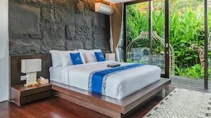A modern bedroom with a stone wall, a large bed with white and blue bedding, a nightstand with a lamp, and a view of lush greenery through a glass door. - De Prestigio Villa II (Canggu)