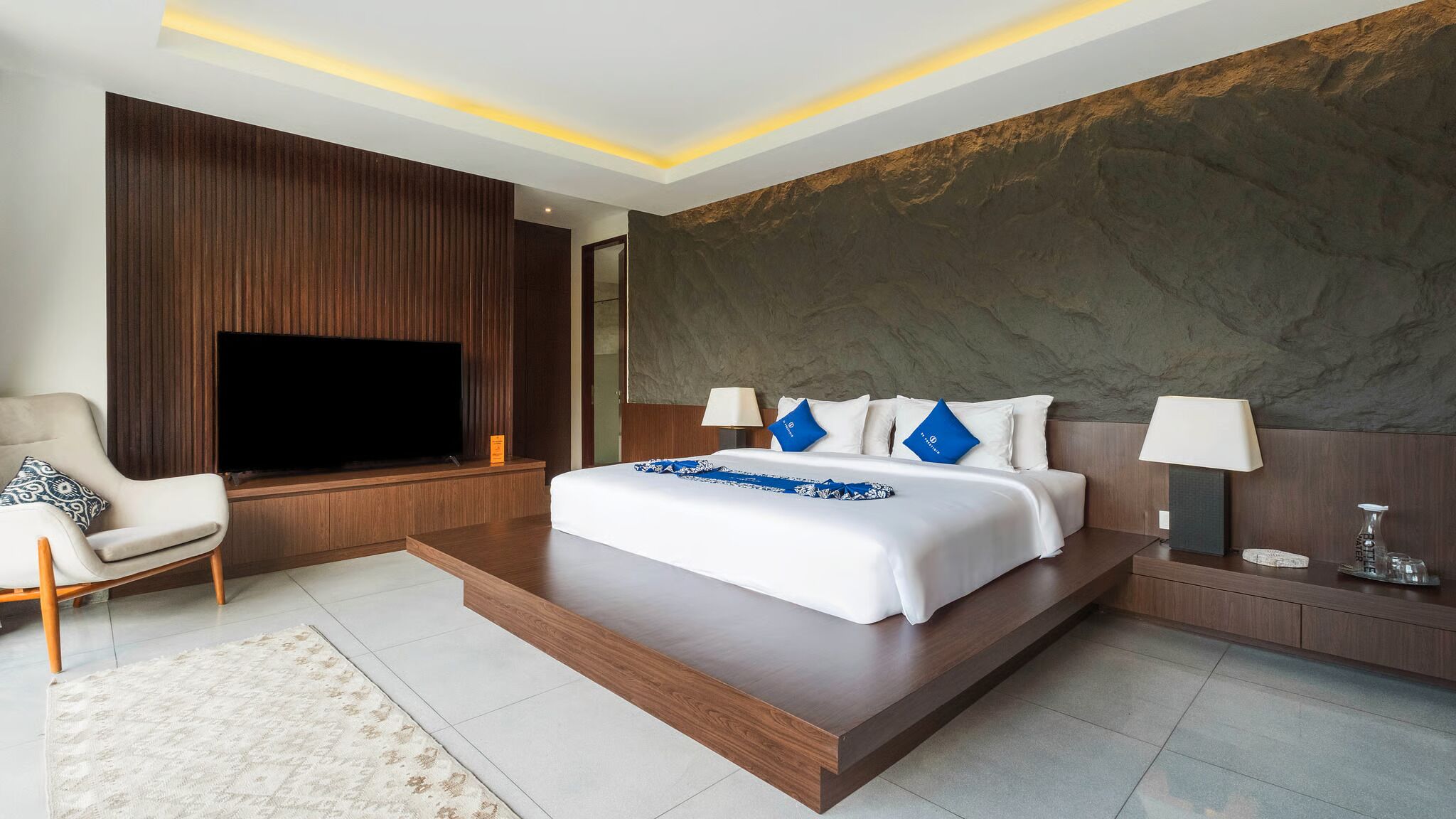 A modern hotel room with a large bed, a flat-screen TV, a seating area with a chair, and a stone accent wall.