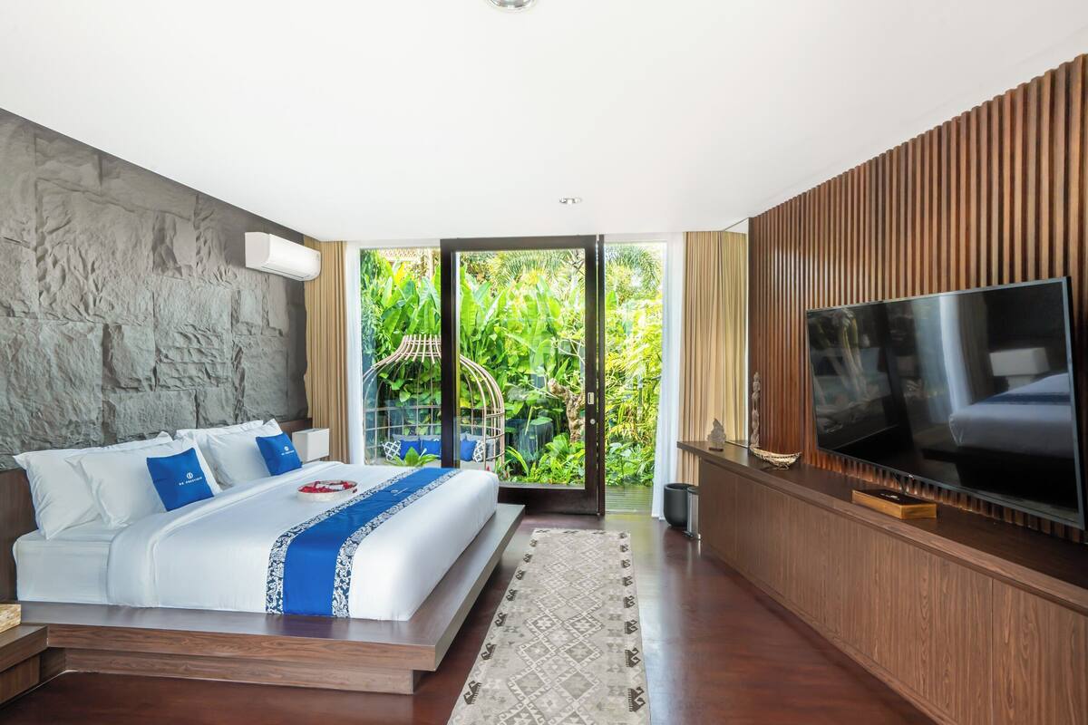 A modern hotel room with a large bed, a flat-screen TV, and a view of lush greenery through the sliding glass door.