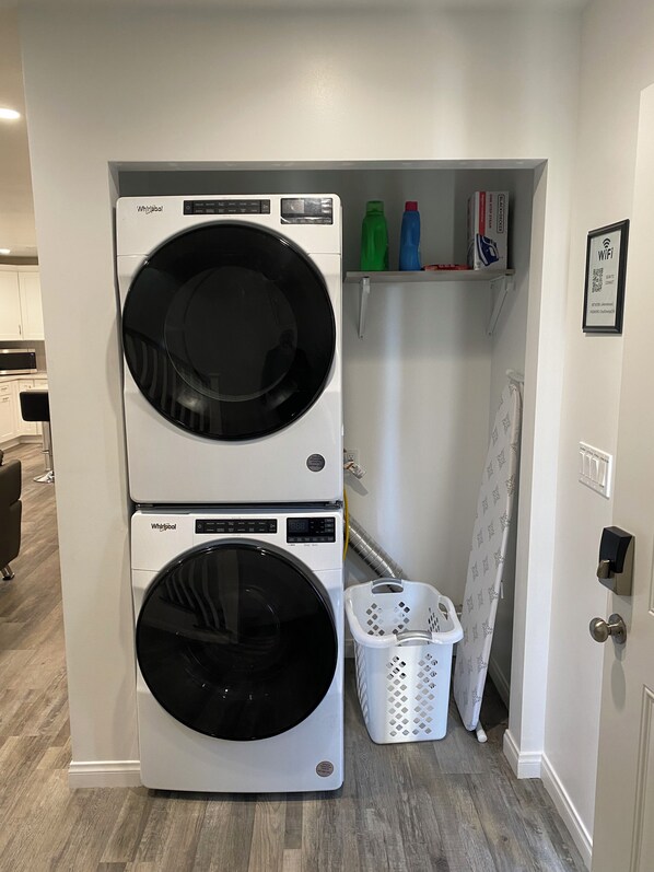 Un lavadero pequeño con dos lavadoras apiladas Whirlpool, una cesta blanca para ropa y un estante con productos de limpieza.