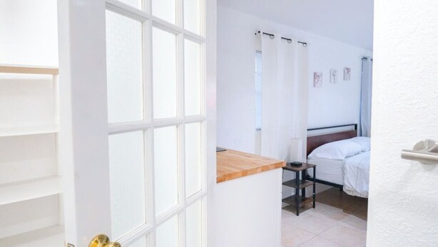 A narrow hallway with a white door leading to a bedroom with a bed, a bedside table, and a window.