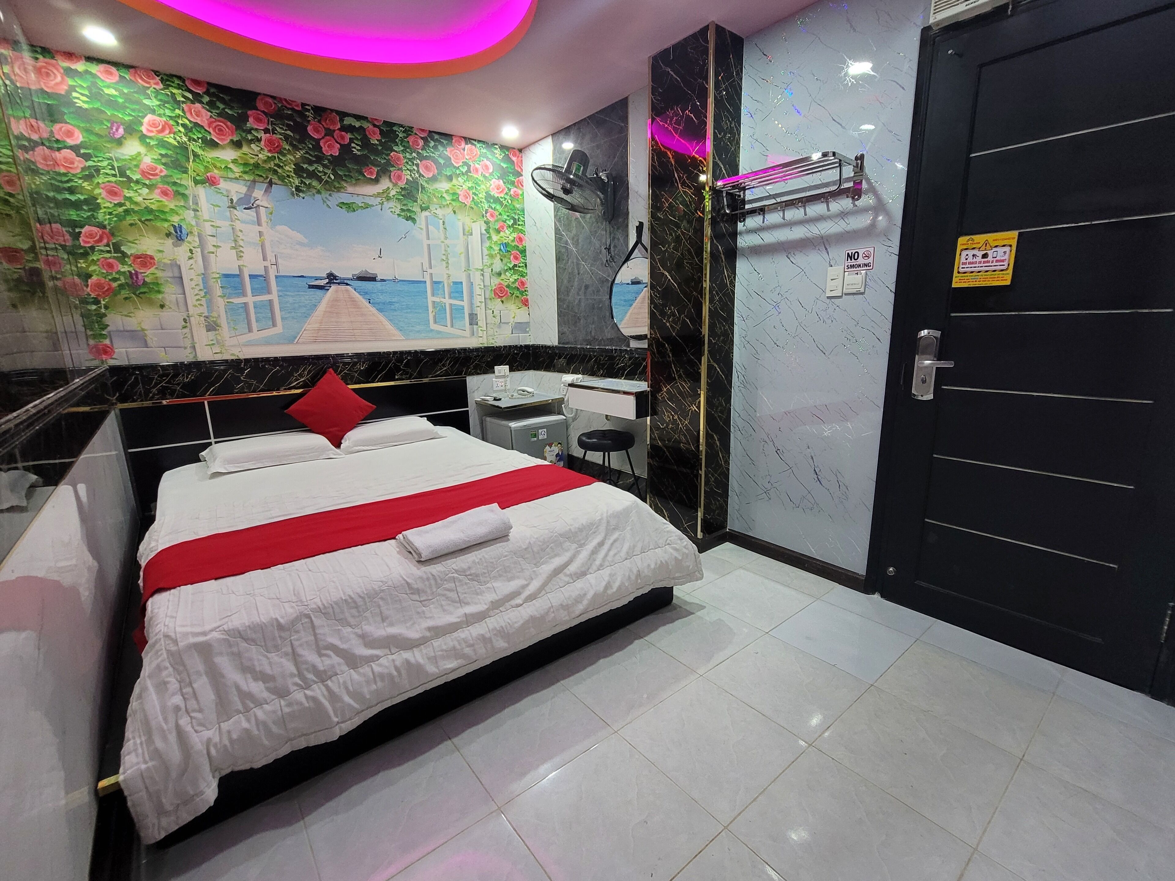 A hotel room with a bed, a desk, a chair, a mirror, a door, and a wall with a floral mural.
