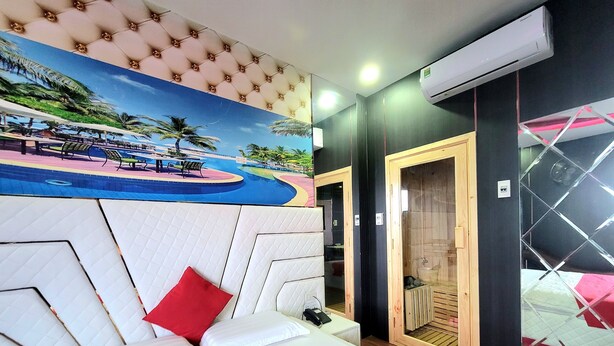 Une chambre d’hôtel avec un grand lit, un oreiller rouge, un tableau représentant un paysage tropical, un appareil de climatisation et une porte en bois.