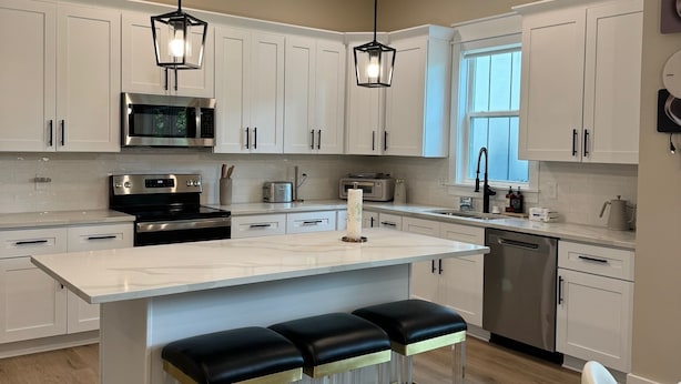 Una cocina moderna con armarios blancos, electrodomésticos de acero inoxidable, una isla central con taburetes altos y luminarias colgantes.