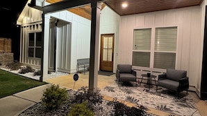 Una casa moderna con un patio cubierto que incluye una mesa y sillas, rodeada de un jardín ajardinado con rocas y arbustos.