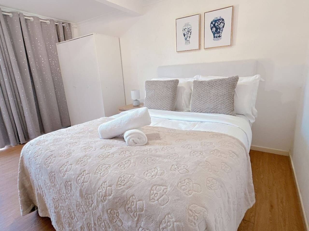 A neatly made bed with a patterned bedspread, a rolled towel, and two framed pictures on the wall.