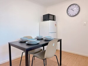 A dining area with a table set for two, a microwave on top of a white refrigerator, and a wall clock.