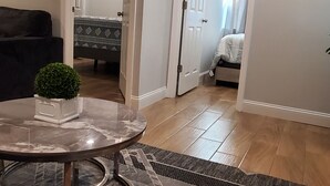 A living room with a round table, a potted plant, and a sofa. There is a door leading to another room with a bed.