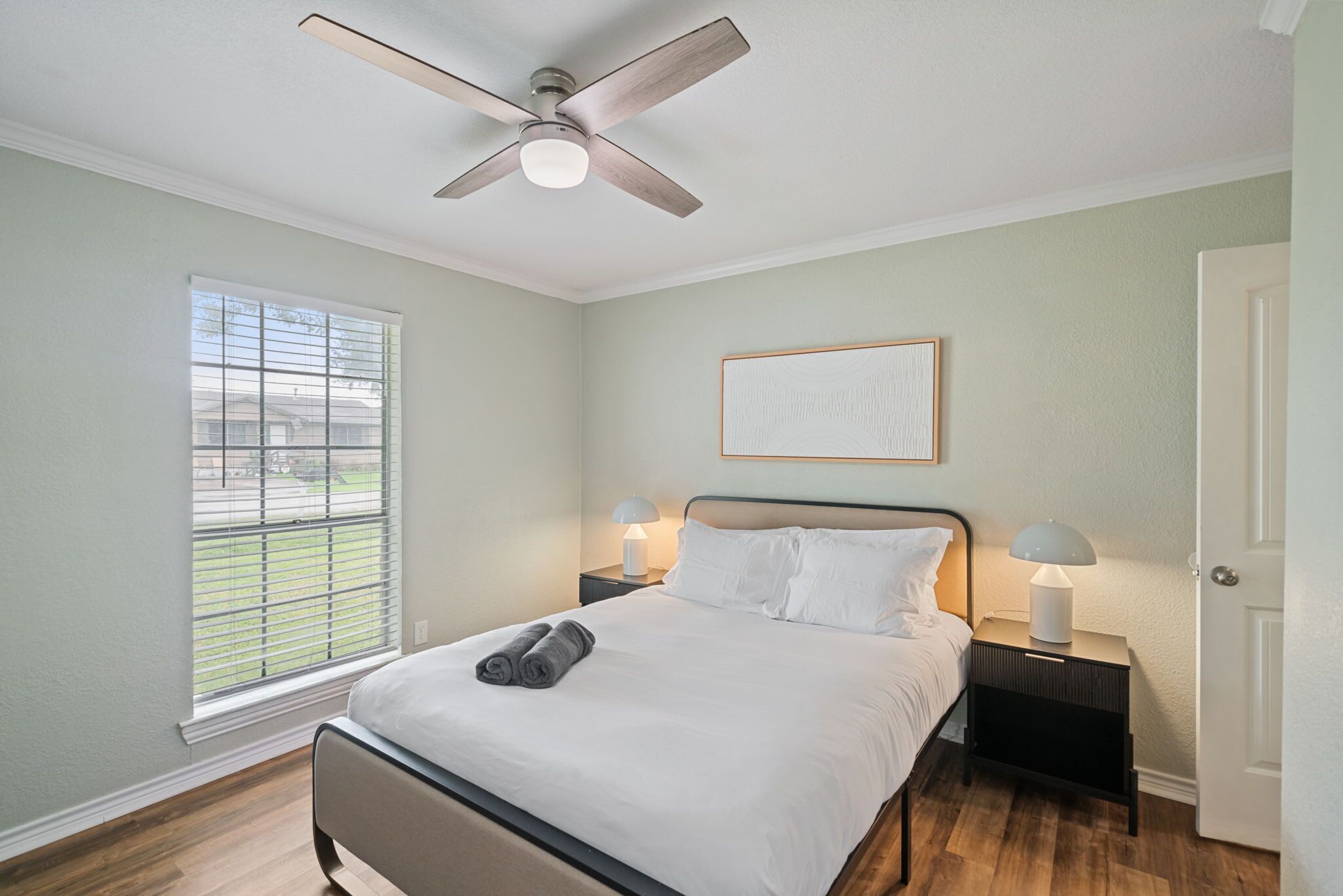 A bedroom with a bed, a ceiling fan, a window with blinds, a bedside table with a lamp, and a door.