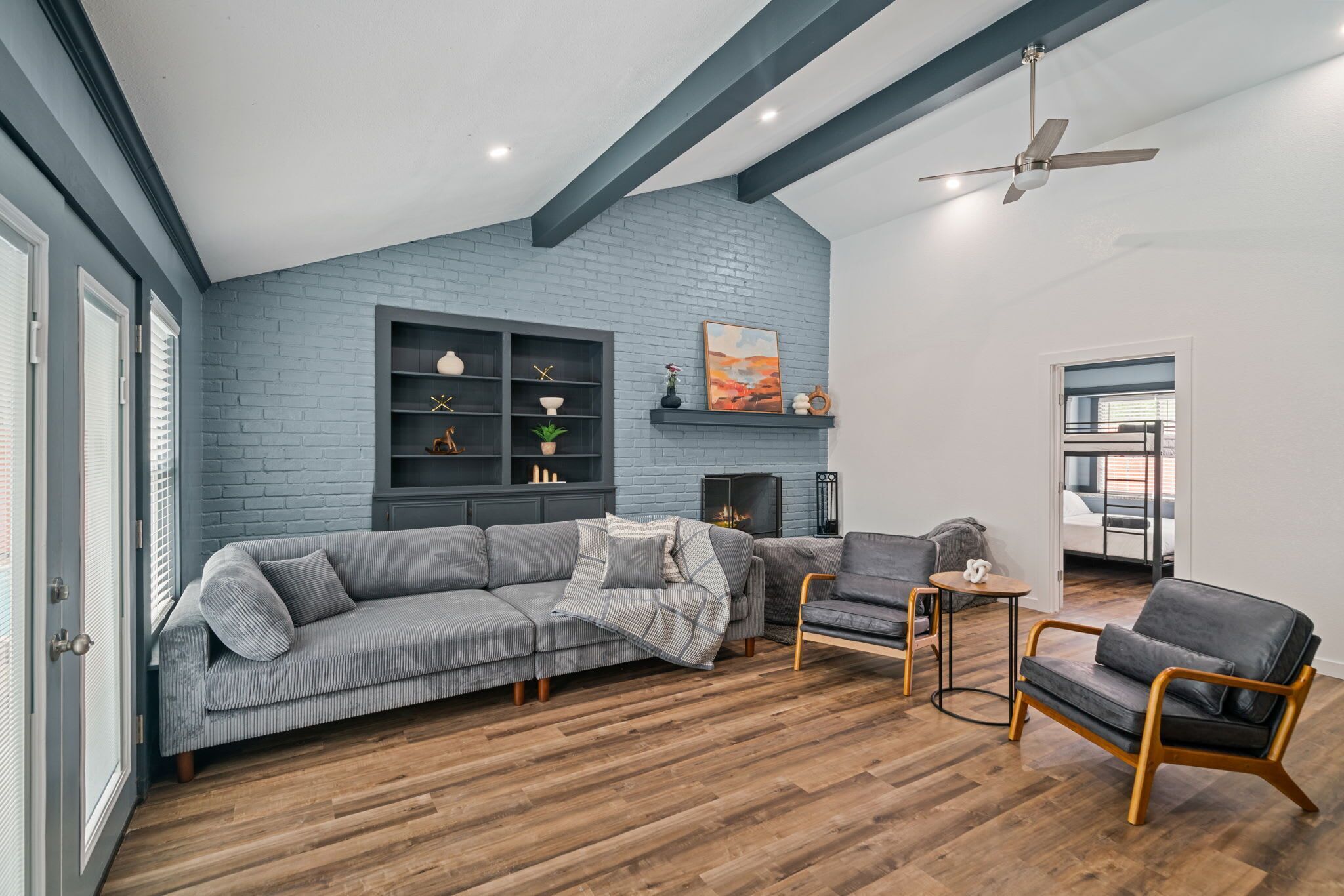 A modern living room with a grey sectional sofa, a small round table, and a grey armchair. There is a fireplace with a mantel, a ceiling fan, and a large window with blinds.