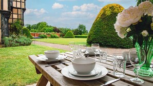 A wooden table set for a meal with white dishes and glasses, a green vase with flowers, and a neatly trimmed hedge in the background. - Severn End - 15th Century Manor House (Worcester)