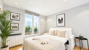 A bedroom with a large bed, a radiator, a window with plants, and framed pictures on the wall.