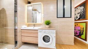 A modern bathroom with a glass shower enclosure, a built-in sink, a washing machine, and two framed artworks on the wall.