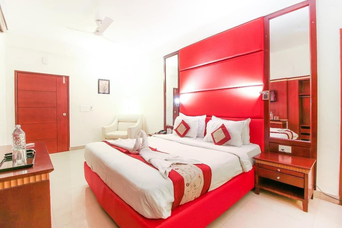 A hotel room with a large bed, a wooden nightstand, a red headboard, and a white chair.