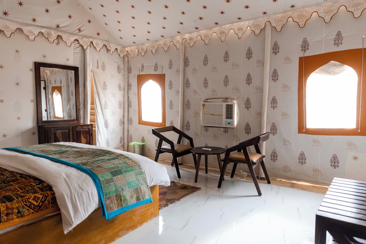 A bedroom with a bed, a desk, two chairs, a mirror, and a window with curtains.