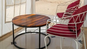 Une petite table ronde en bois, dotée de pieds en métal, deux chaises en métal blanc avec des coussins rouges, et une grande fenêtre en arc de cercle offrant une vue sur un paysage désertique.