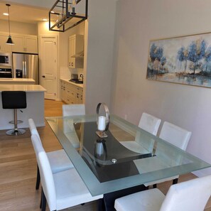 A modern dining area with a glass-top table, white upholstered chairs, and a painting on the wall.