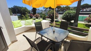 A patio with a glass table and chairs, a yellow umbrella, and a view of a tennis court and greenery.