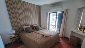 A bedroom with a bed, a bedside table, a lamp, a window with curtains, and a wall-mounted air conditioner.