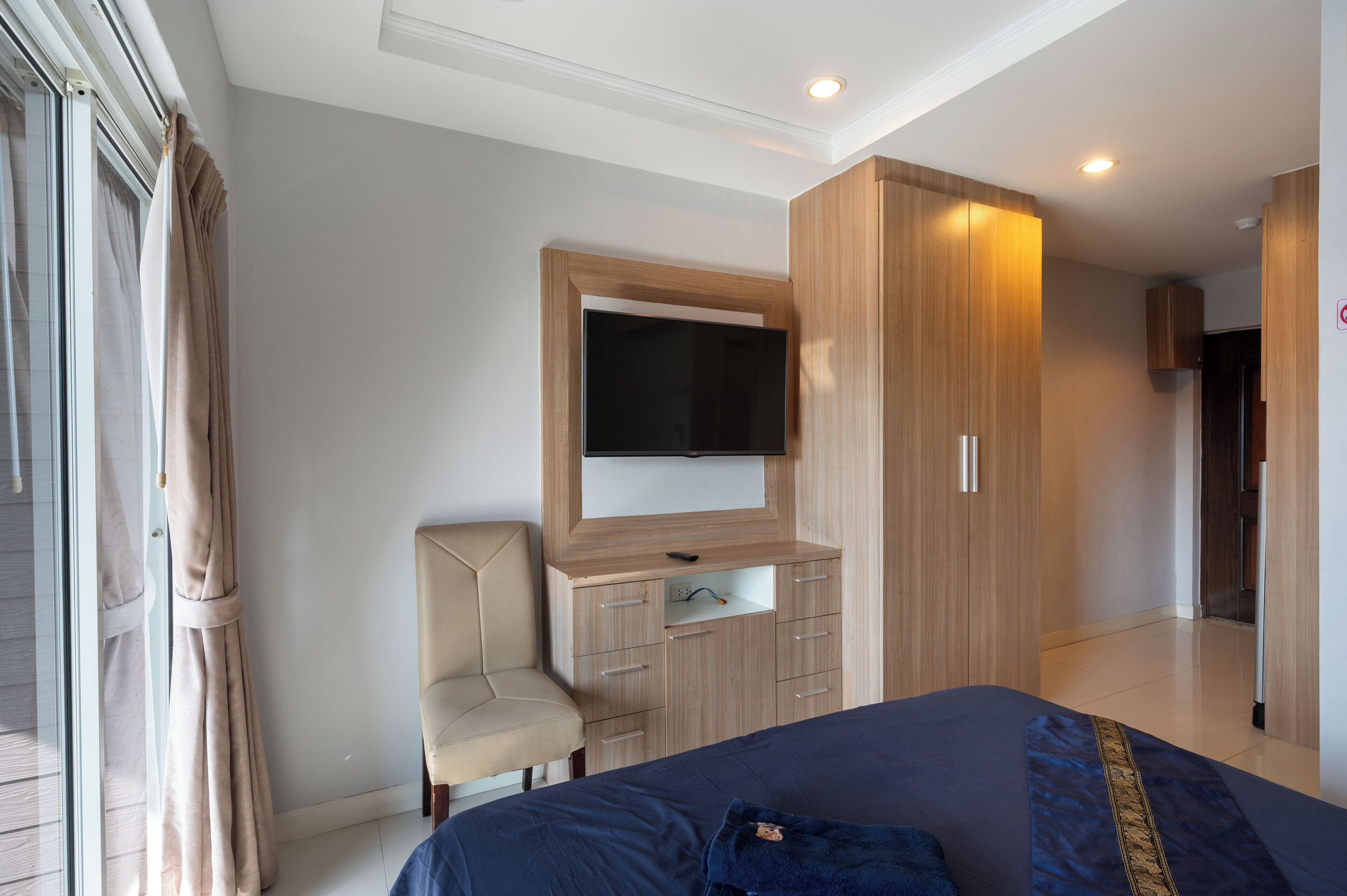 Une chambre à coucher comprenant un lit, un téléviseur fixé sur un meuble en bois, une chaise et une porte-fenêtre coulissante.