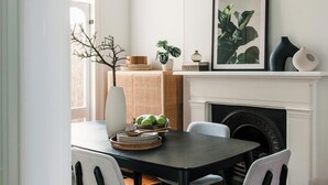 Un coin repas avec une table noire, des chaises blanches, une cheminée et une plante encadrée accrochée au mur.