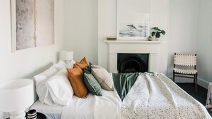 Une chambre à coucher avec un lit, des oreillers, une lampe de chevet, une cheminée, une chaise et un tableau au mur.