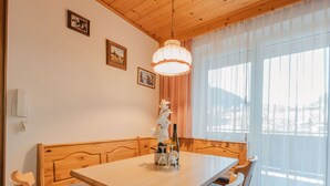 A dining area with a wooden table and chairs, a chandelier, and a view of a snowy landscape through large windows. - Beautiful apartment for 4 people with TV and balcony (Neukirchen Am Großvenediger)
