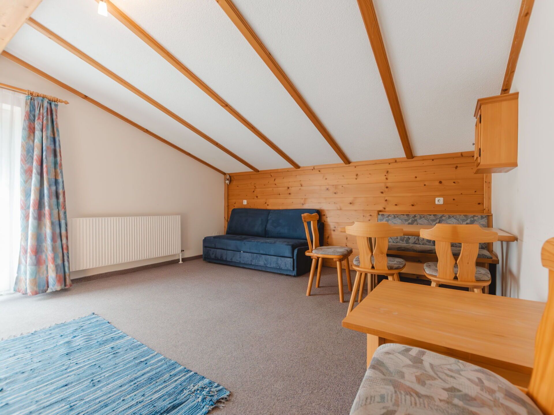 A cozy attic room with a wooden ceiling, a blue sofa, a wooden dining table, and a radiator.