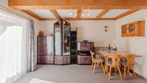 A room with a wooden ceiling, a dining table with chairs, a cabinet, and a window with curtains.