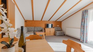 A cozy attic room with a wooden ceiling, a sofa, a TV, and a dining table with a bottle of wine.