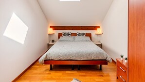 Une chambre avec un plafond en pente, un puits de lumière, une tête de lit en bois, des lampes de chevet, un lit avec une literie à motifs, une commode en bois et une petite plante en pot.