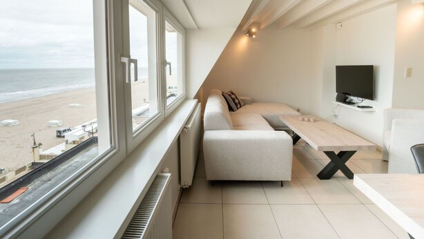 A modern room with a large window overlooking a beach, a sofa, a TV, and a table. - Lovely apartment for 4 guests with WIFI and TV (De Haan)