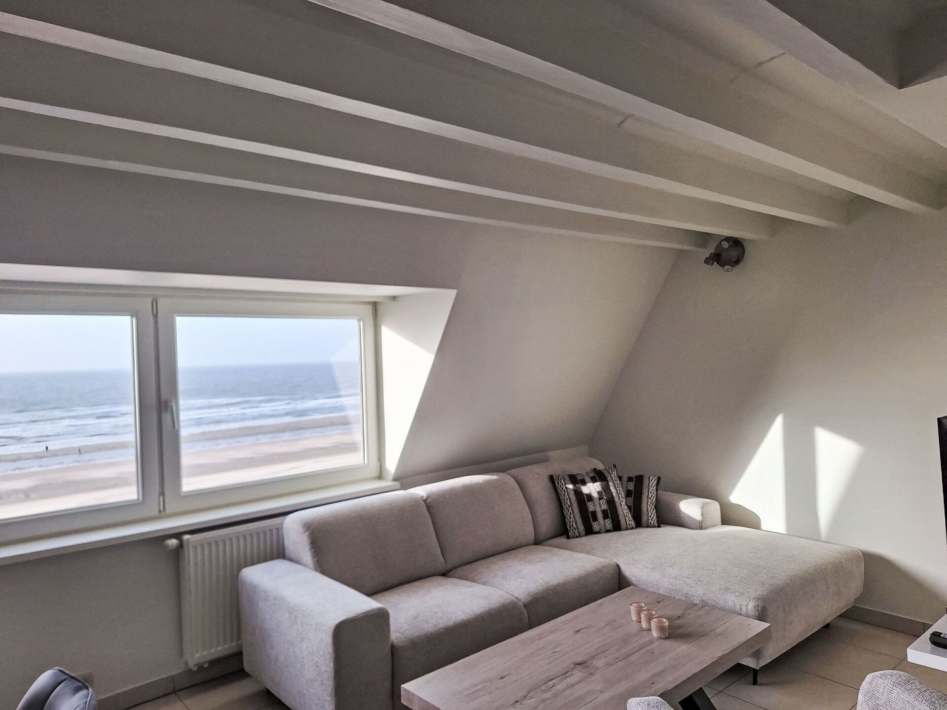 Una sala de estar moderna con un ventanal grande que da a la playa, un sofá gris, una mesa de centro de madera y un radiador.