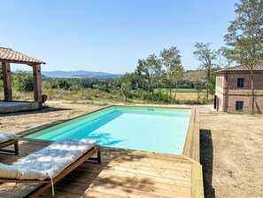 Ein rechteckiger Swimmingpool mit Holzterrasse und Liegestühlen, umgeben von ländlicher Landschaft mit Bäumen und einem fernen Gebirgszug.