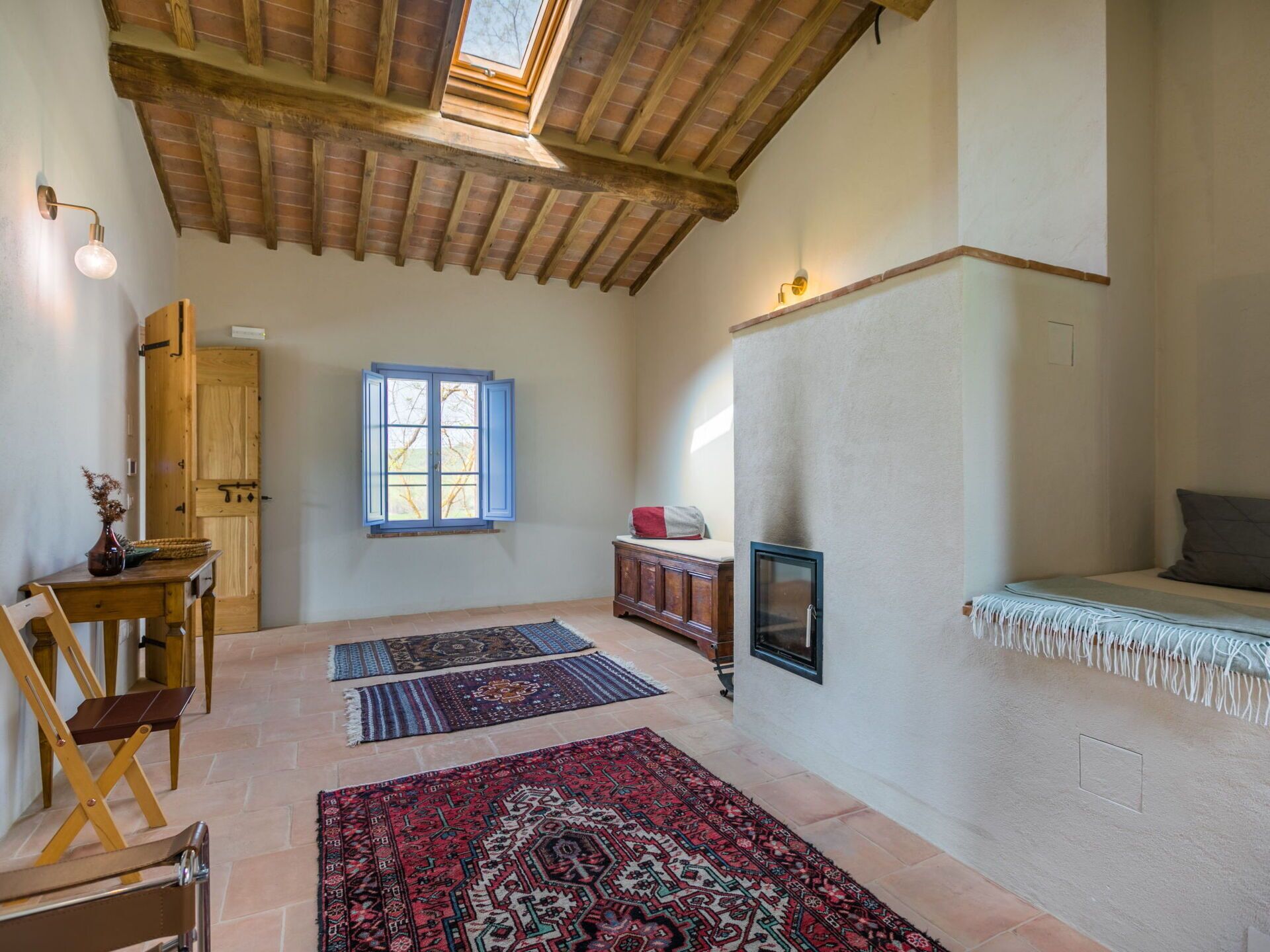 Une pièce spacieuse avec un plafond haut, une grande fenêtre et une cheminée. Plusieurs tapis sont posés sur le sol, et on y trouve une table en bois avec des chaises.