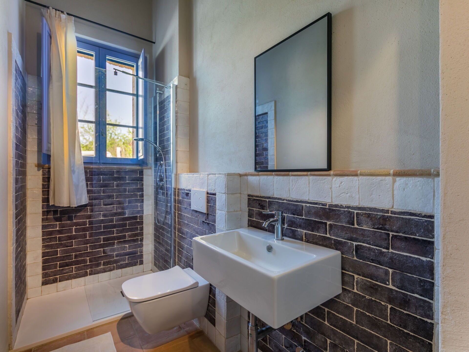A bathroom with a shower, toilet, and sink, featuring a brick wall and a window with curtains.