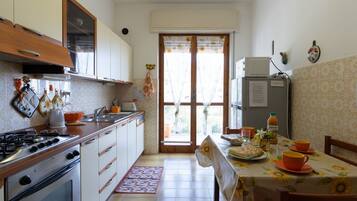 Une cuisine compacte équipée d’une cuisinière à gaz, d’un four et d’un réfrigérateur. On y trouve une table à manger recouverte d’une nappe à motifs floraux et agrémentée de tasses orange.
