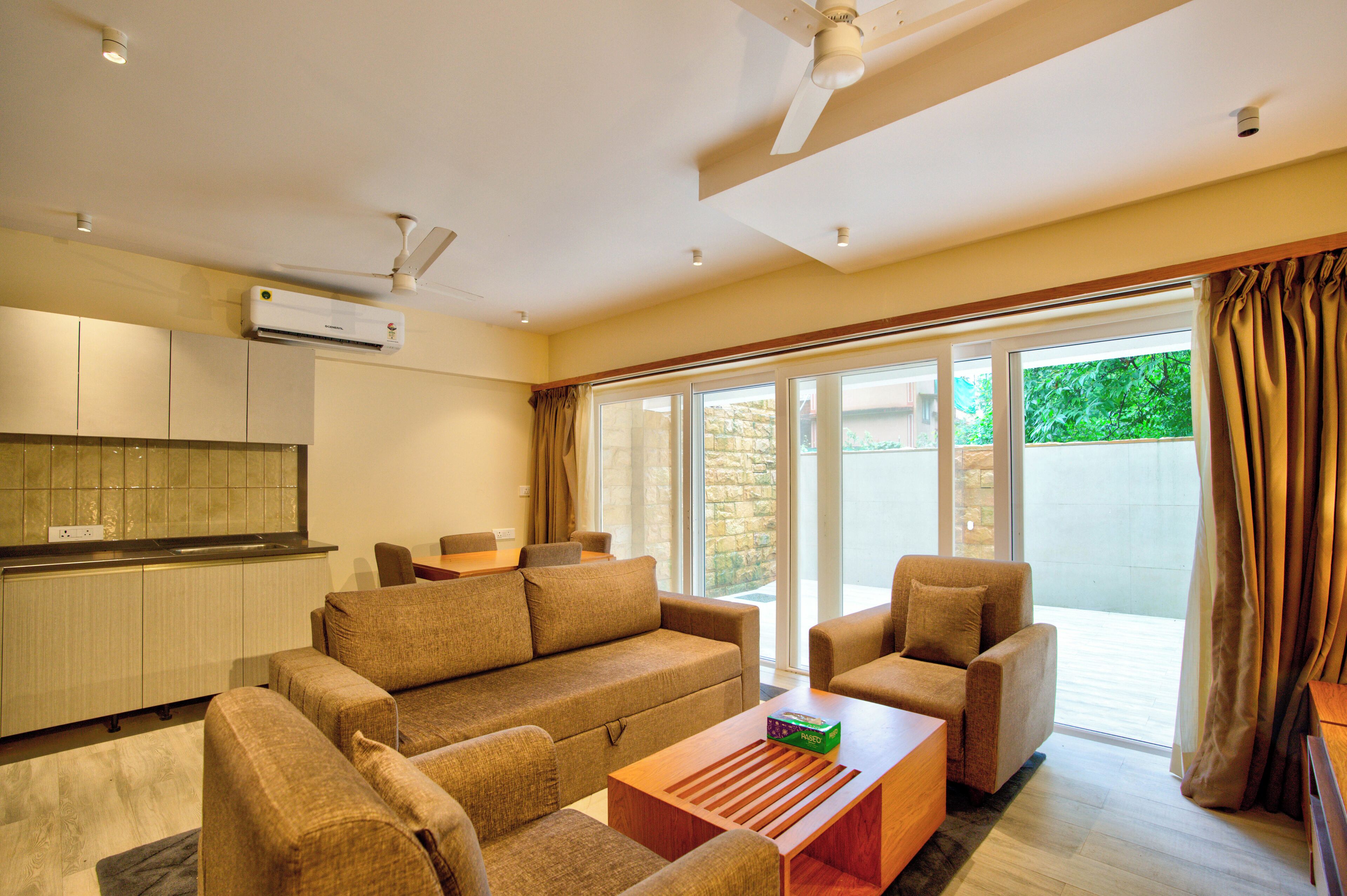 A modern living room with a sofa, armchairs, and a coffee table. There is a kitchen area with cabinets and an air conditioning unit. Large sliding glass doors lead to an outdoor area.
