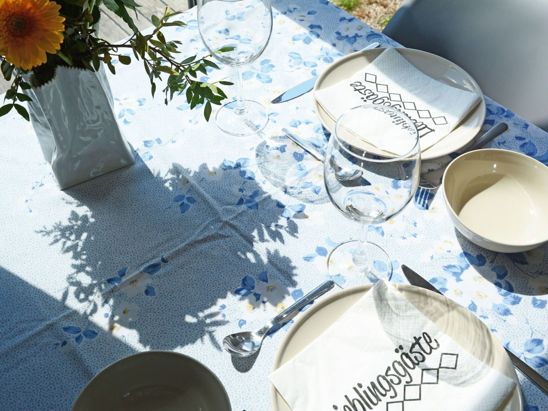 A table set with plates, a glass, a bowl, and a spoon, with a floral tablecloth and a vase with a flower.