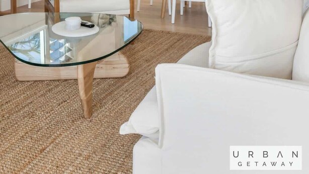 Un interior moderno con un sofá blanco, una mesa de centro de vidrio, una zona de comedor con sillas blancas, un ventilador de techo y una cocina con armarios blancos.