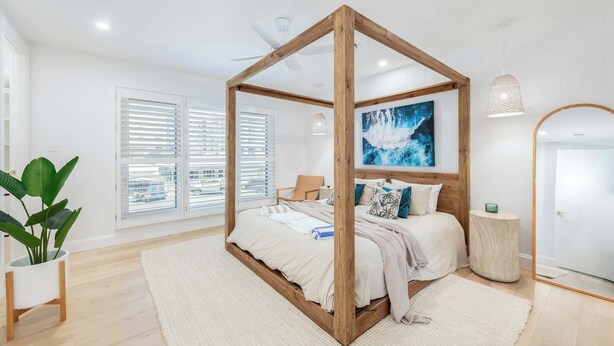 A bedroom with a four-poster bed, a large window with blinds, a potted plant, and a painting on the wall.