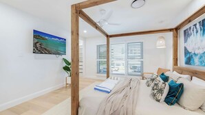 A bedroom with a bed, a ceiling fan, a window with blinds, a painting on the wall, and a plant.