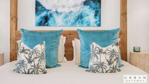 Un dormitorio con un cuadro de una gran ola marina, dos almohadas a juego y un cabecero de madera.