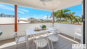 A covered outdoor area with a table and chairs, a ceiling fan, and a view of palm trees and buildings.