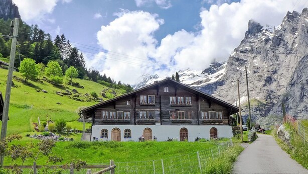 Un chalet traditionnel, doté d’une façade en bois, de nombreuses fenêtres et de fondations en pierre, qui se détache sur un paysage montagneux et un ciel dégagé.