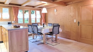 Un intérieur en bois comprenant une cuisine et un coin repas, avec un îlot central, des chaises, une table et une horloge au mur.