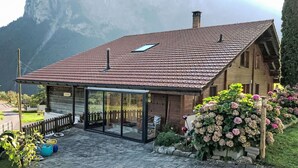 Un chalet avec un toit recouvert de tuiles rouges, une véranda fermée par des vitres et un jardin agrémenté d’hortensias.